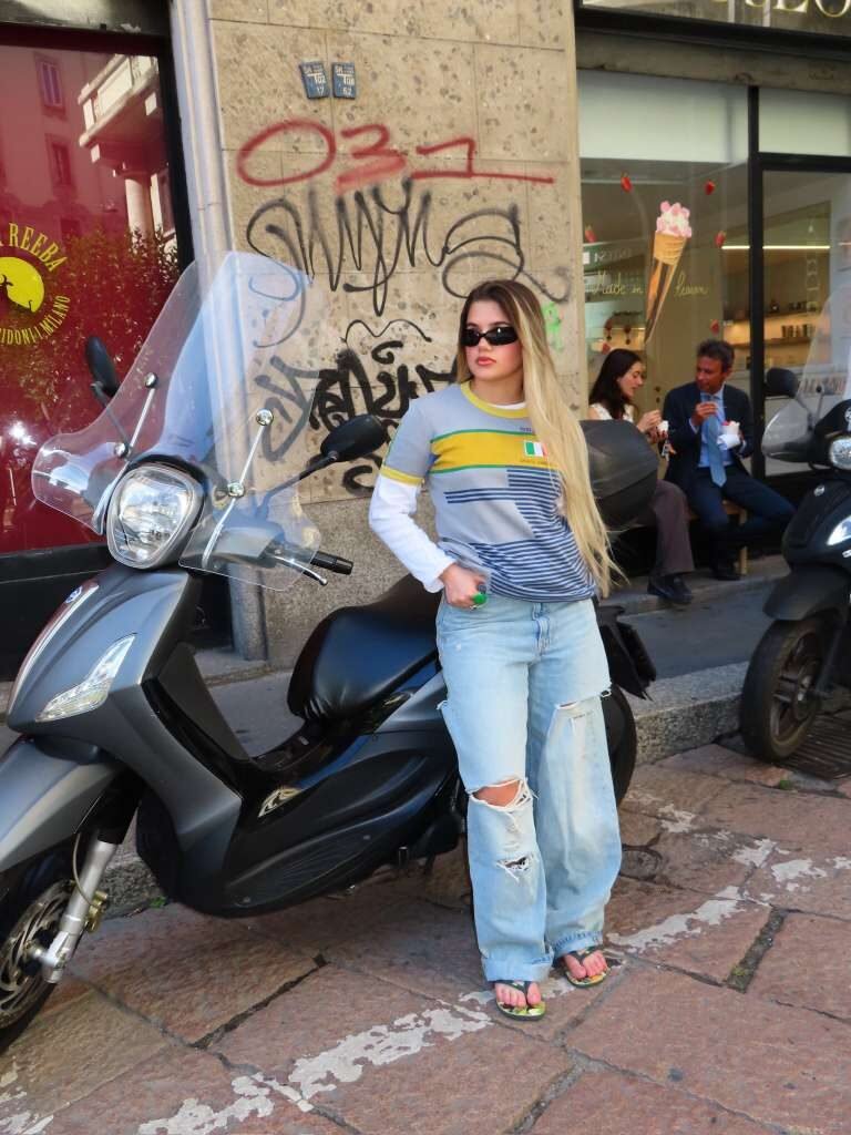 Young woman in a vintage striped tee and ripped wide-leg jeans leaning on a scooter on a graffitied street.