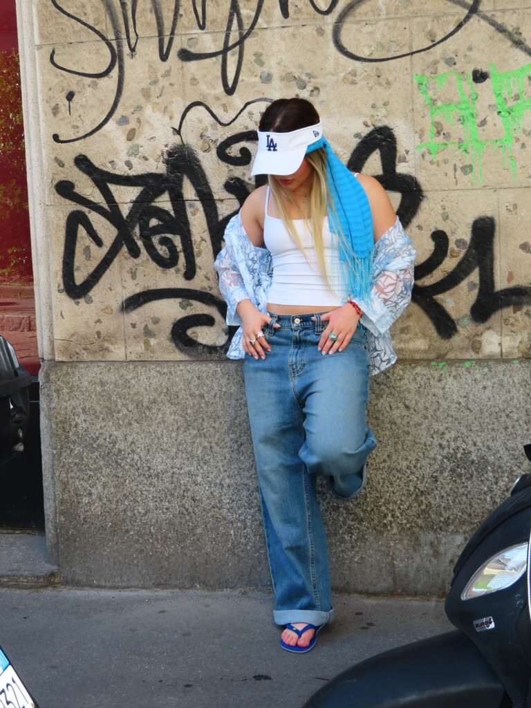 Young woman in an LA visor, white tank and baggy jeans against a graffiti wall.