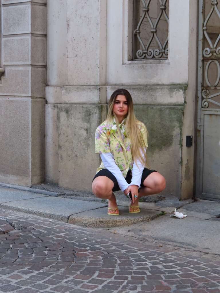 Young woman crouched on a stone step wearing a floral second-hand shirt over a white long sleeve.