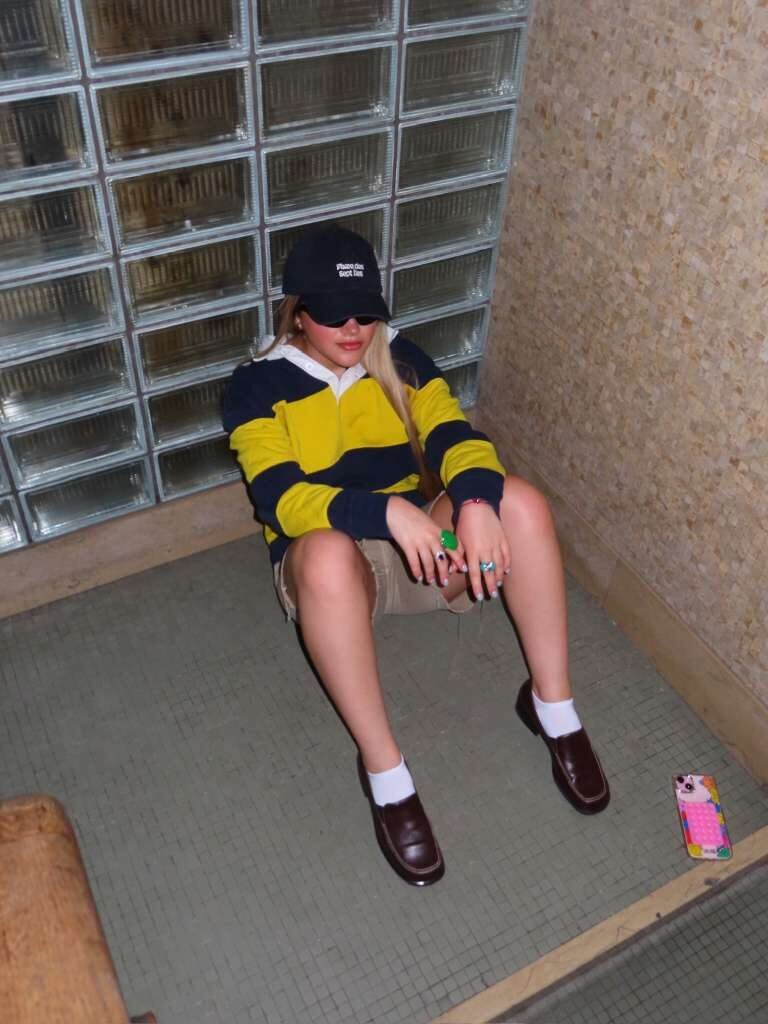 Young woman seated wearing a striped rugby polo, cap, white socks and brown loafers.