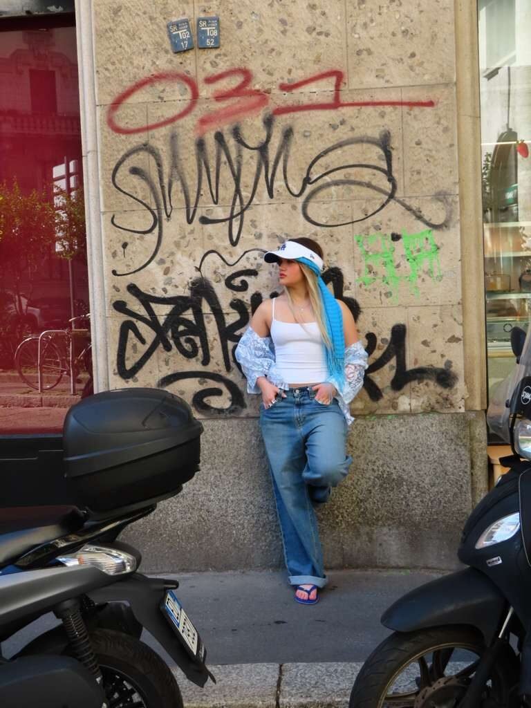 Wide street shot of a young woman in baggy jeans and a visor, framed by parked scooters.