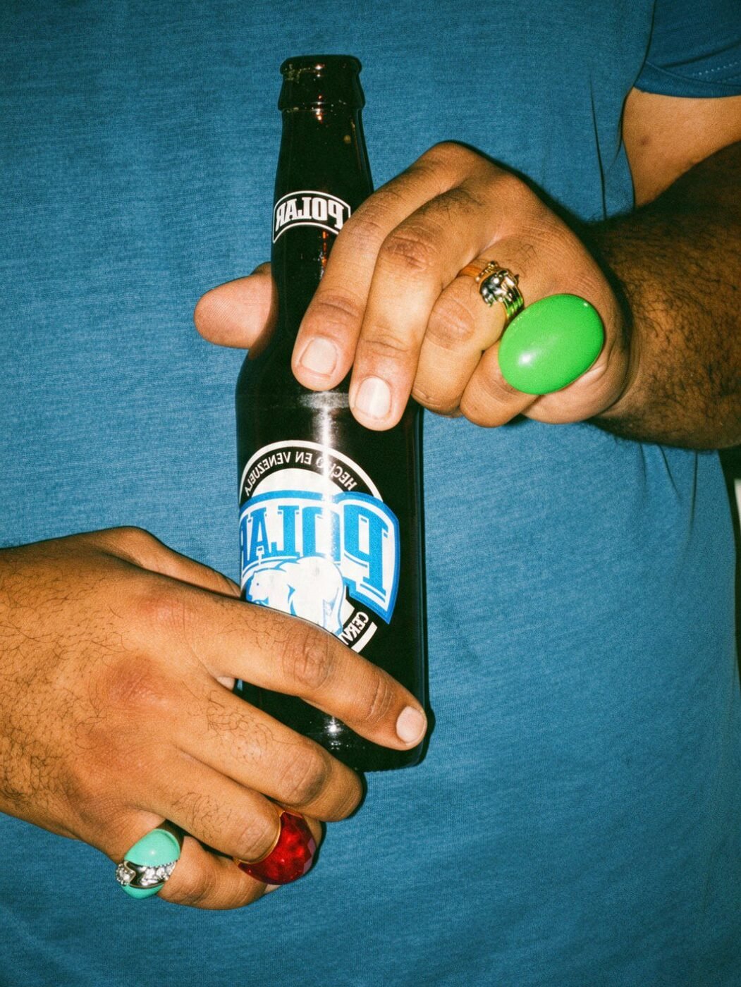 Close-up of two hands with a green stone ring and gold signet rings holding a cold Polar beer bottle.