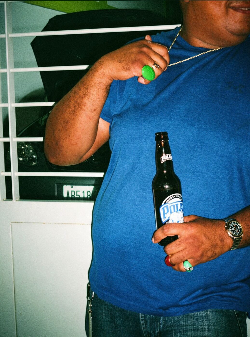 Man in a blue tee with a gold rope chain and rings holding a Polar beer beside a car at night.