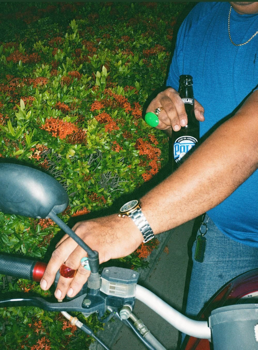 Night shot of a man with a Polar beer beside a motorbike, framed by orange ixora flowers.