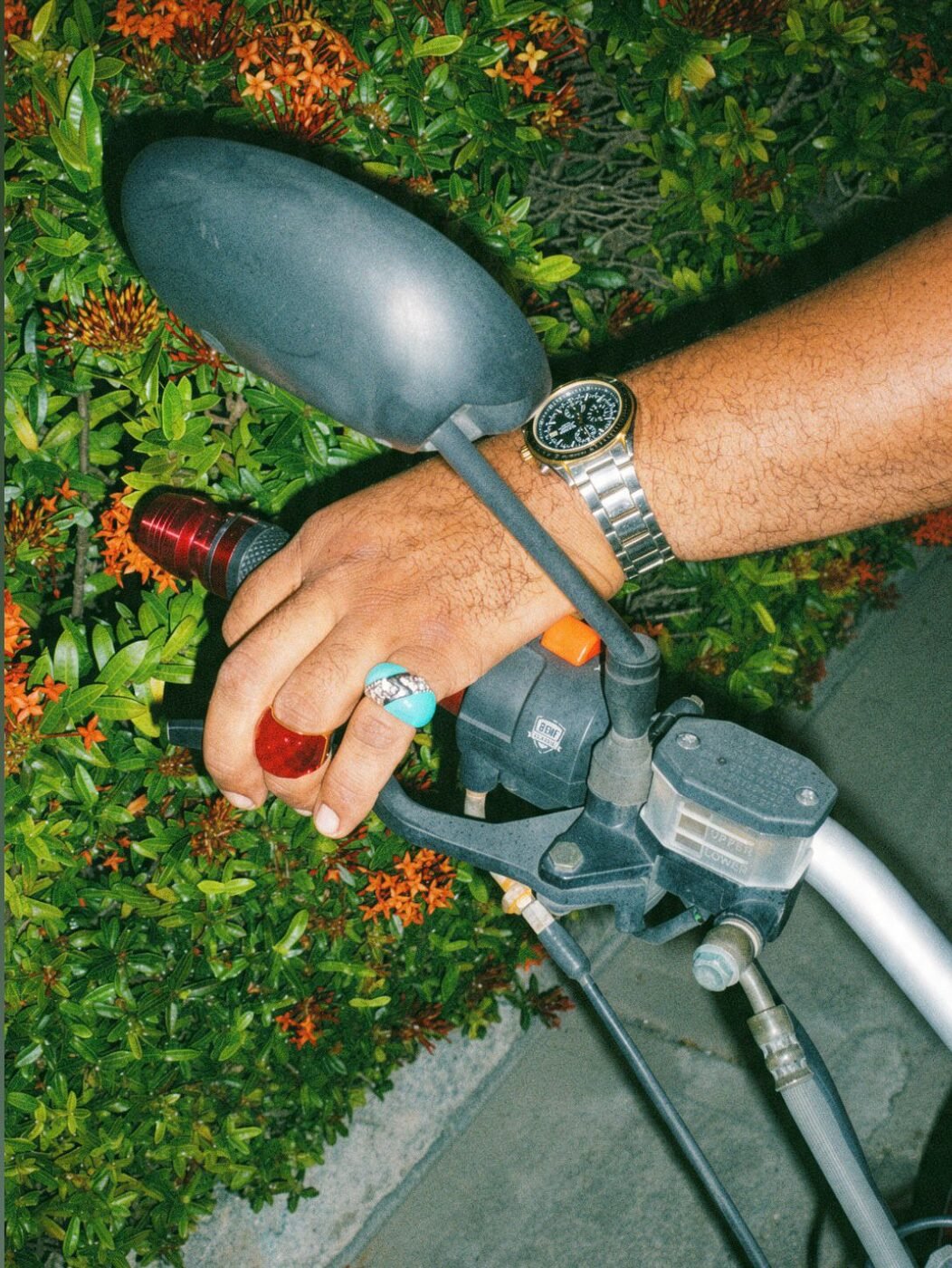 Hand resting on motorbike handlebars wearing turquoise and red rings and a steel watch.